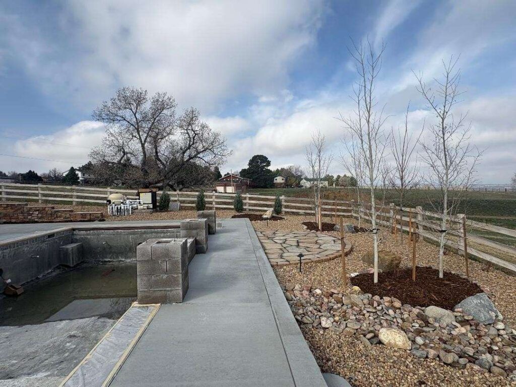 Concrete walkway leading toward a stone pond with landscaping, young trees, and a wooden fence under a cloudy sky.
