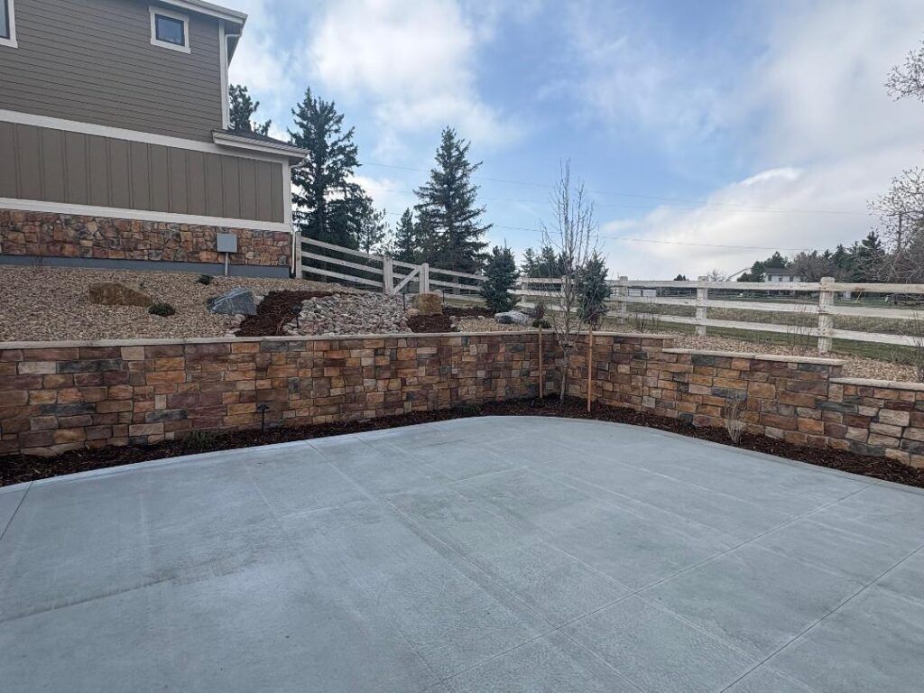 A new concrete patio with a tiered stone retaining wall, mulch beds, and a white fence against a cloudy blue sky.