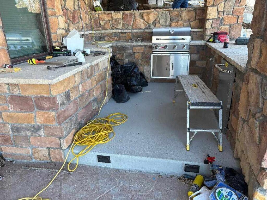 An outdoor kitchen area with stone counters, a stainless steel grill, a yellow extension cord, and a silver work bench.