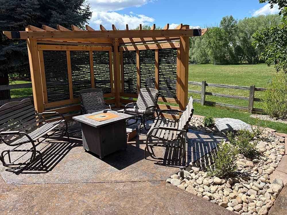 A patio with a fire pit, wooden pergola, and garden chairs, situated on a stone floor next to a green lawn and fence.