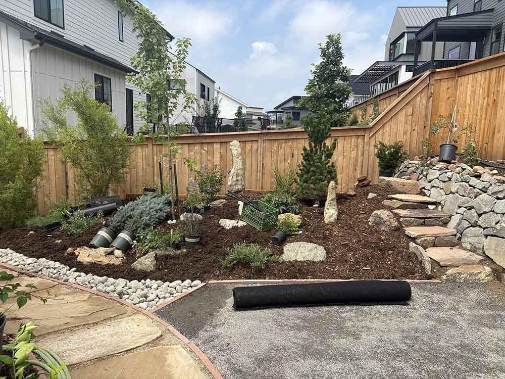 A backyard garden with mulch, rock features, and small plants under a wooden fence, with stone steps along the side.