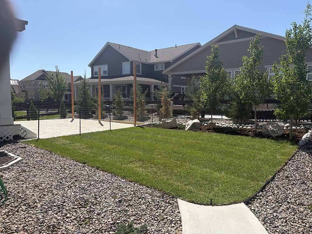 A sunny backyard with a green lawn, gravel border, concrete patio, and wooden posts under a clear blue sky.