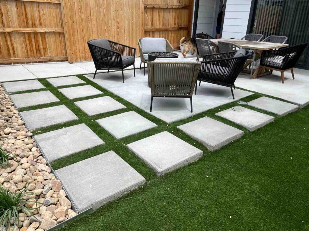 A modern backyard patio features gray square pavers set in grass, a concrete sitting area, outdoor furniture, and rocks.
