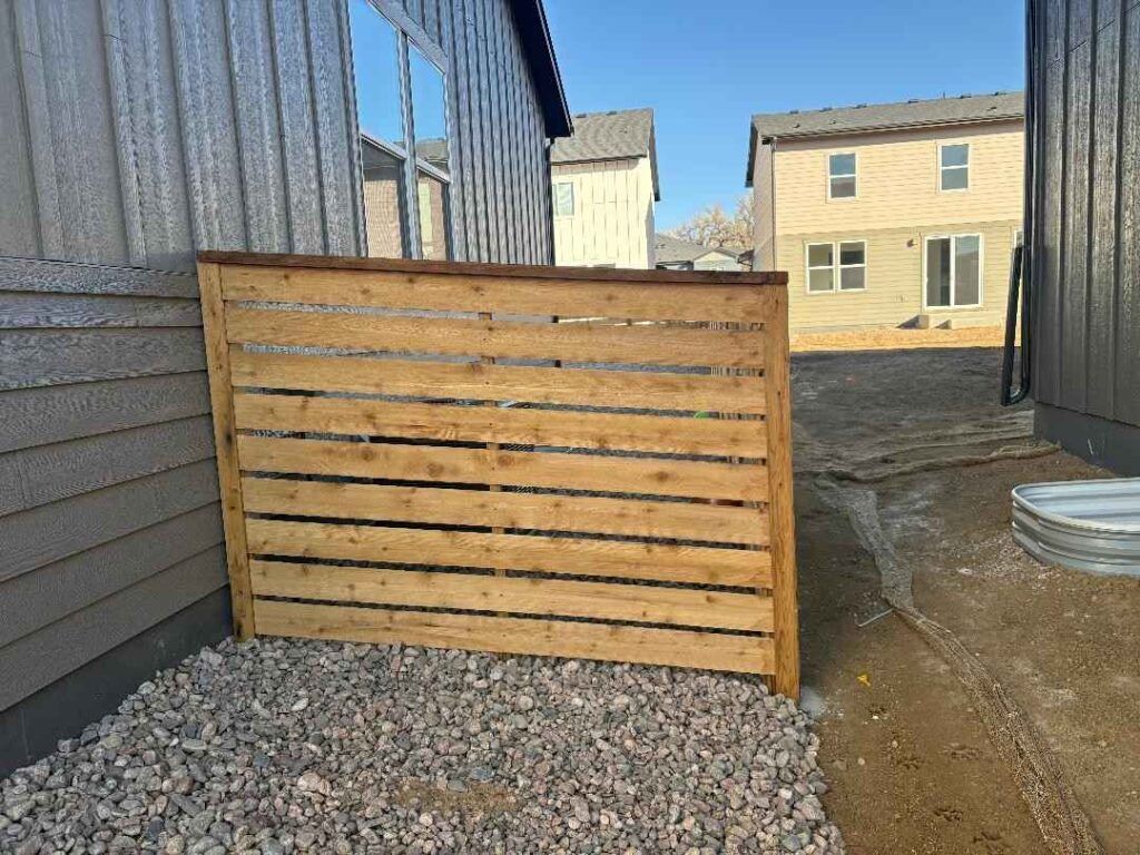 A wood horizontal slat privacy screen sits on a gravel yard between two house walls with an unfinished home in the distance.
