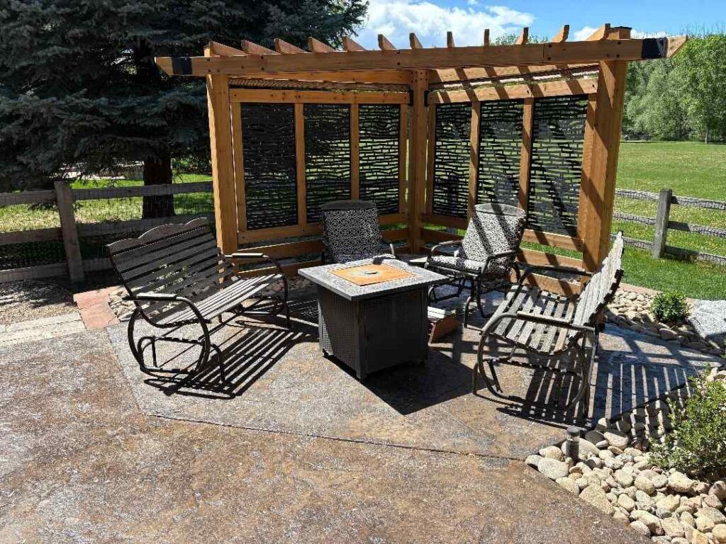 A wooden pergola covers a patio seating area with benches, chairs, and a central square fire pit on a sunny day.