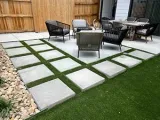 A backyard patio featuring square concrete pavers set in artificial grass, with outdoor seating arranged on a deck.