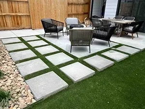 A backyard patio featuring square concrete pavers set in artificial grass, with outdoor seating arranged on a deck.