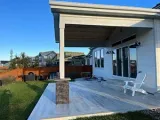 A white-sided house with a covered stone-pillared patio, light gray wood-look flooring, and a single white Adirondack chair.