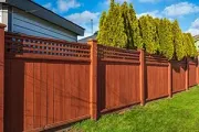 A reddish-brown wooden fence with a lattice top, set against a green lawn and tall evergreen trees under a blue sky.