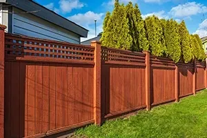 A reddish-brown wooden fence with a lattice top, set against a green lawn and tall evergreen trees under a blue sky.