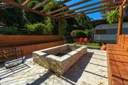 A stone fire pit surrounded by built-in wooden bench seating on a patio under a wooden pergola, near a parked camper.