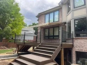 A view of a modern brown wood deck with stairs and a black metal railing attached to the back of a brick house.