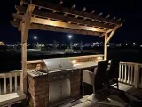 An outdoor kitchen with a stainless steel grill and stone base sits under a wooden pergola on a deck at night.