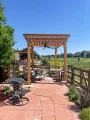A stone patio with outdoor seating under a wooden pergola, overlooking a grassy field under a clear blue sky.