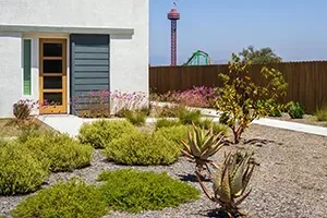 A backyard patio featuring square concrete pavers set in artificial grass, with outdoor seating arranged on a deck.