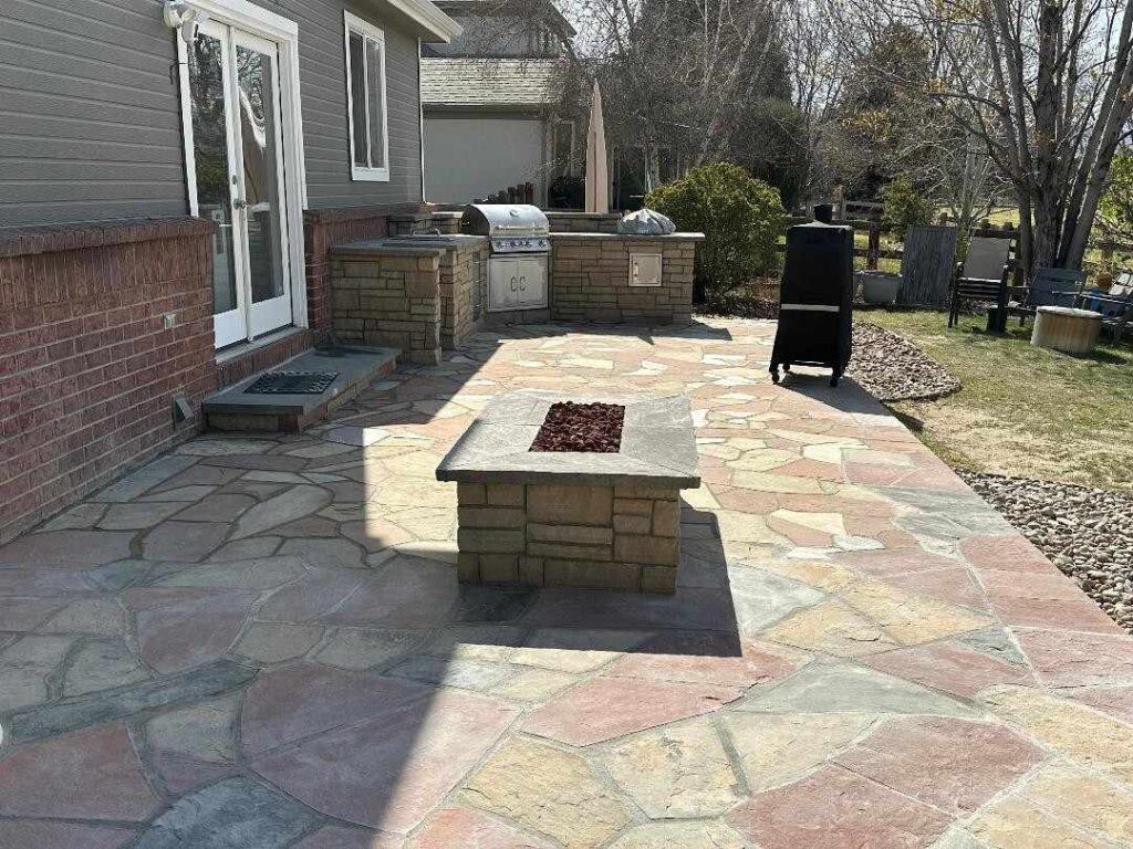 A stone patio featuring a central fire pit, an outdoor kitchen with a grill, and a brick house exterior.
