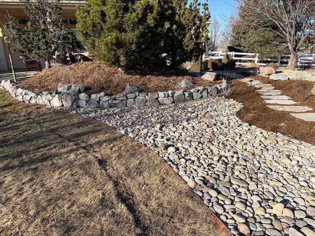 A landscape scene features a dry creek bed of grey stones, a stone retaining wall, mulch, and a stone garden path.