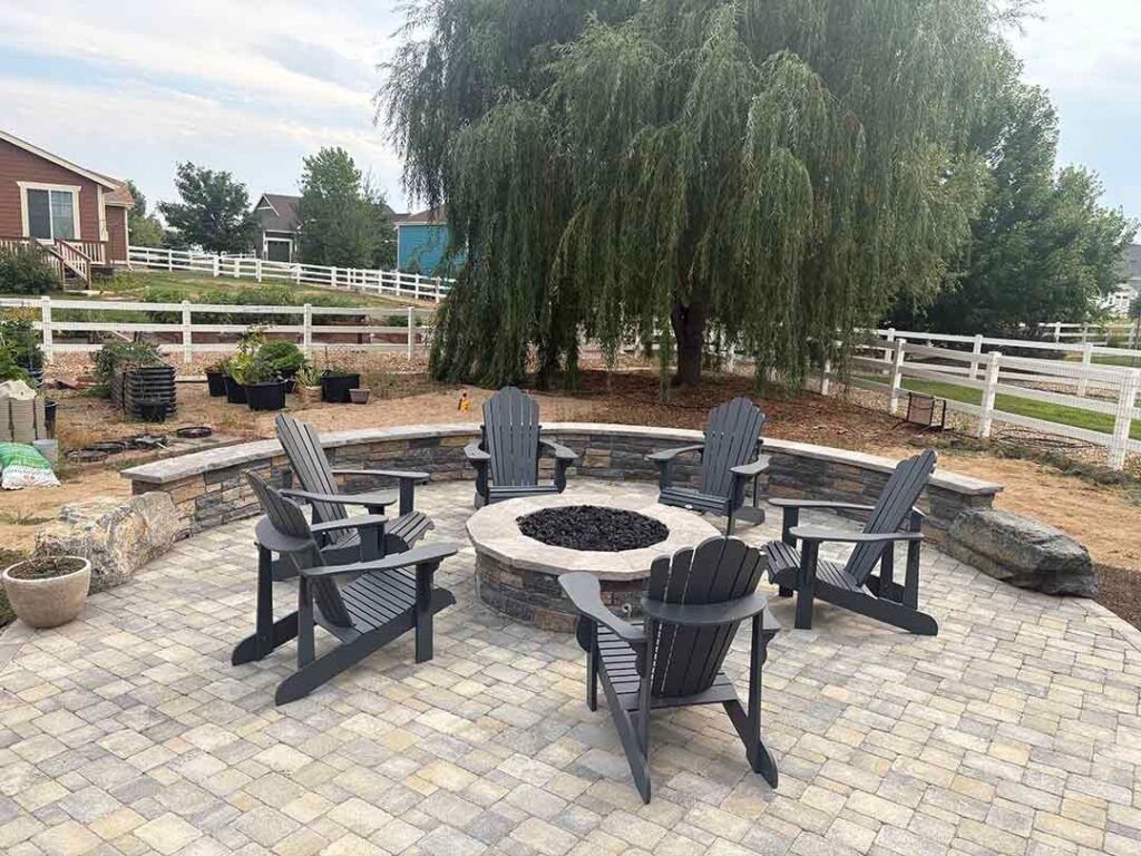 Five dark grey Adirondack chairs arranged in a circle around a stone fire pit on a paved backyard patio.
