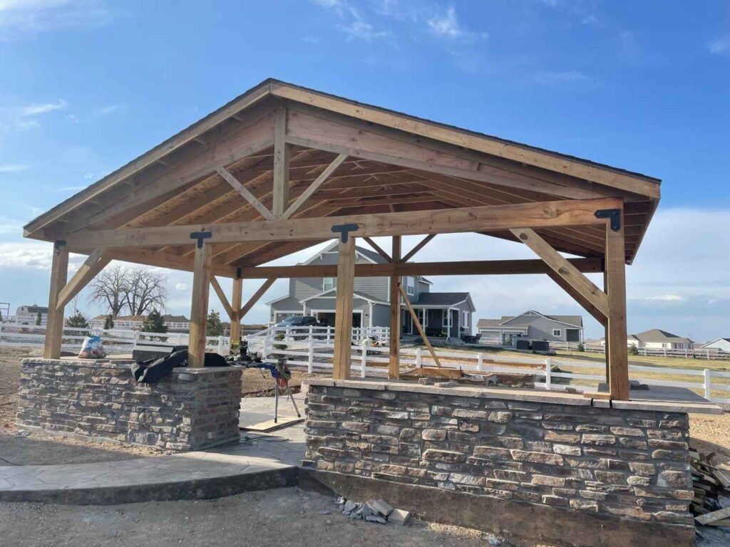 An outdoor wooden pavilion with a stone base under a blue sky, set in a backyard near a house.