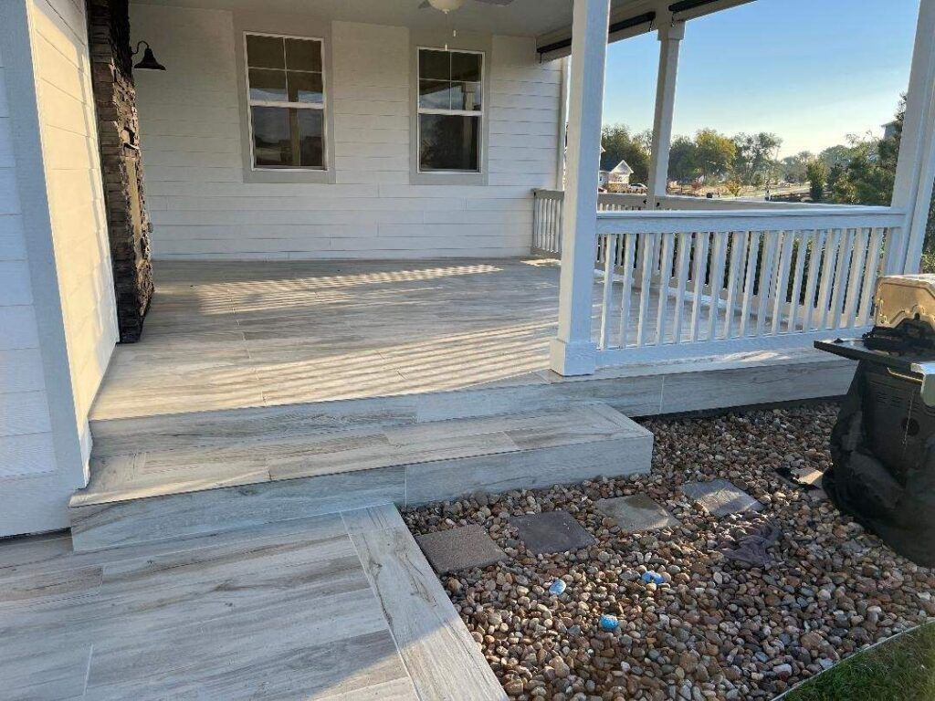 A white porch with light wood-look floor tiles and a railing, featuring a stone step leading to a graveled yard.