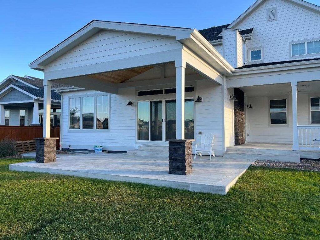 A white house with a covered stone patio, featuring large sliding glass doors and a grassy yard under a clear blue sky.