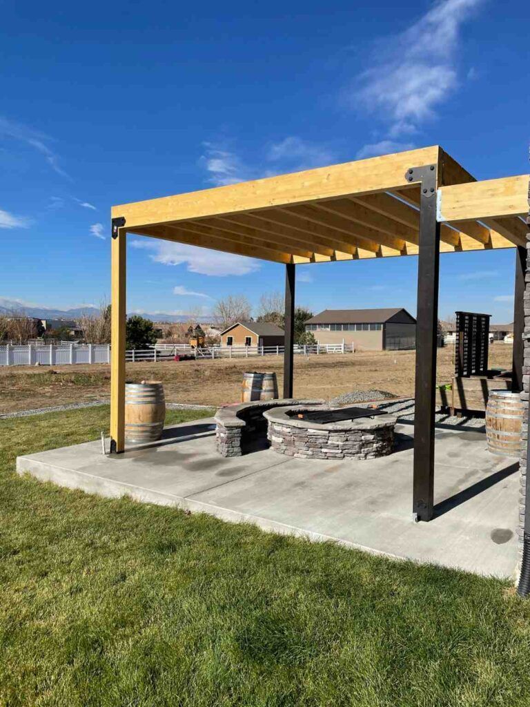 A wooden pergola over a stone fire pit on a concrete patio in a grassy yard under a clear blue sky.