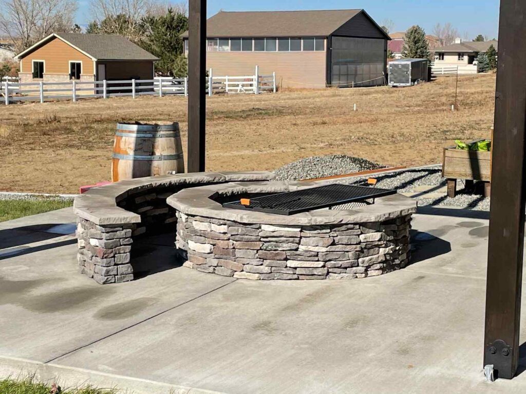 Stone fire pit with a metal grate on a concrete patio, with barrels and rural buildings in the background.