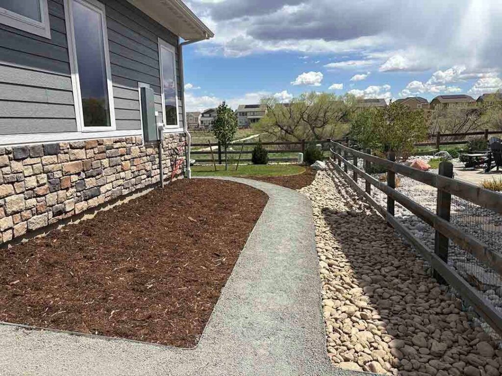 A gravel path separates a mulched garden bed next to a stone-accented house from a wooden fence bordering a grassy field.