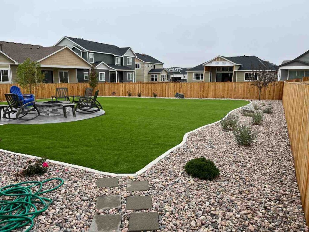 A landscaped backyard featuring an artificial turf lawn, a stone patio with chairs, and gravel beds bordered by a fence.