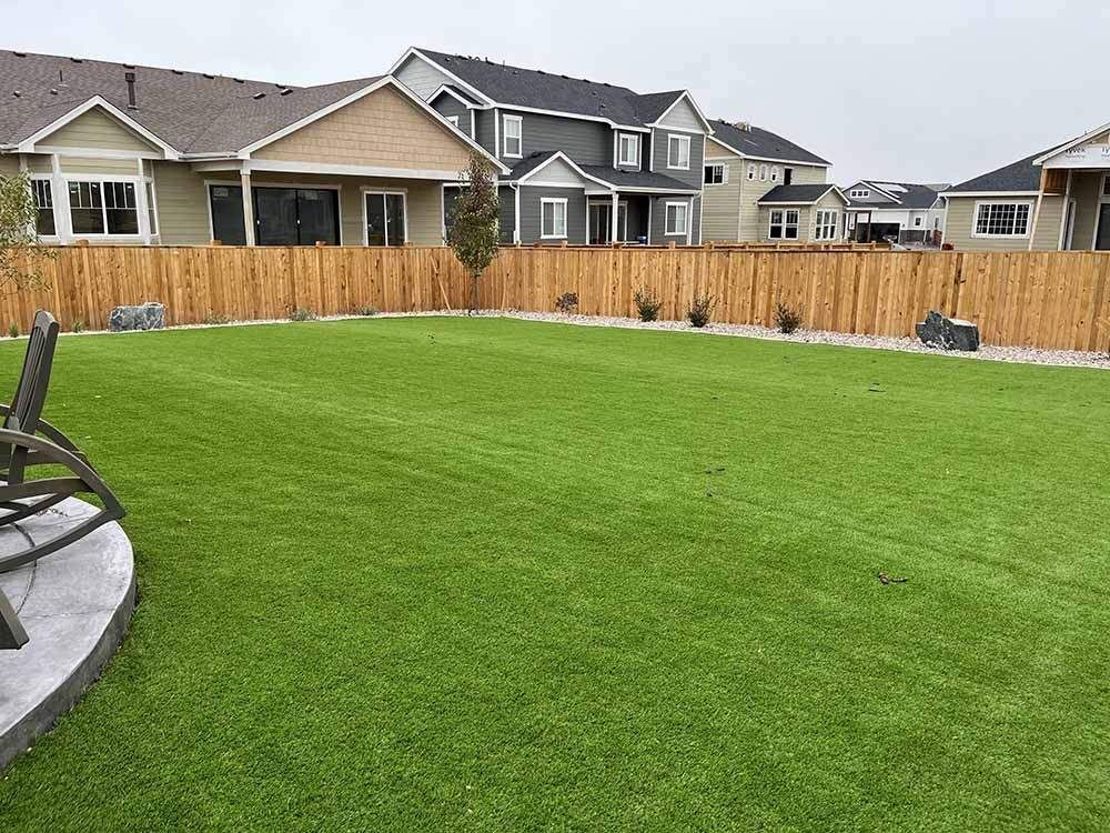 A green backyard lawn bordered by a wood fence with houses in the background and a portion of a patio chair on the left.
