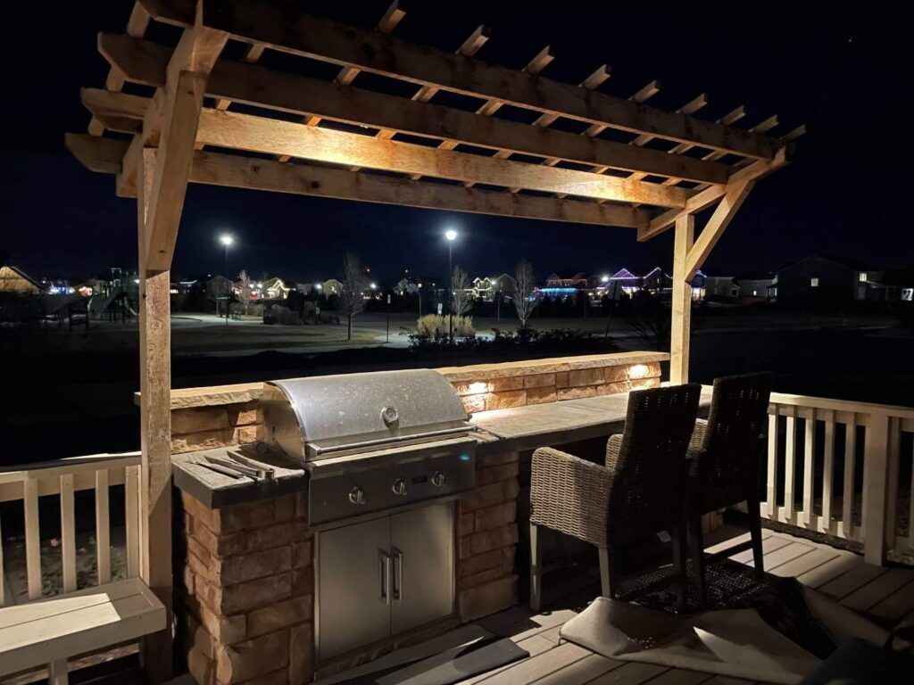 An outdoor stone kitchen island with a stainless steel grill under a wooden pergola on a deck at night, facing a town.