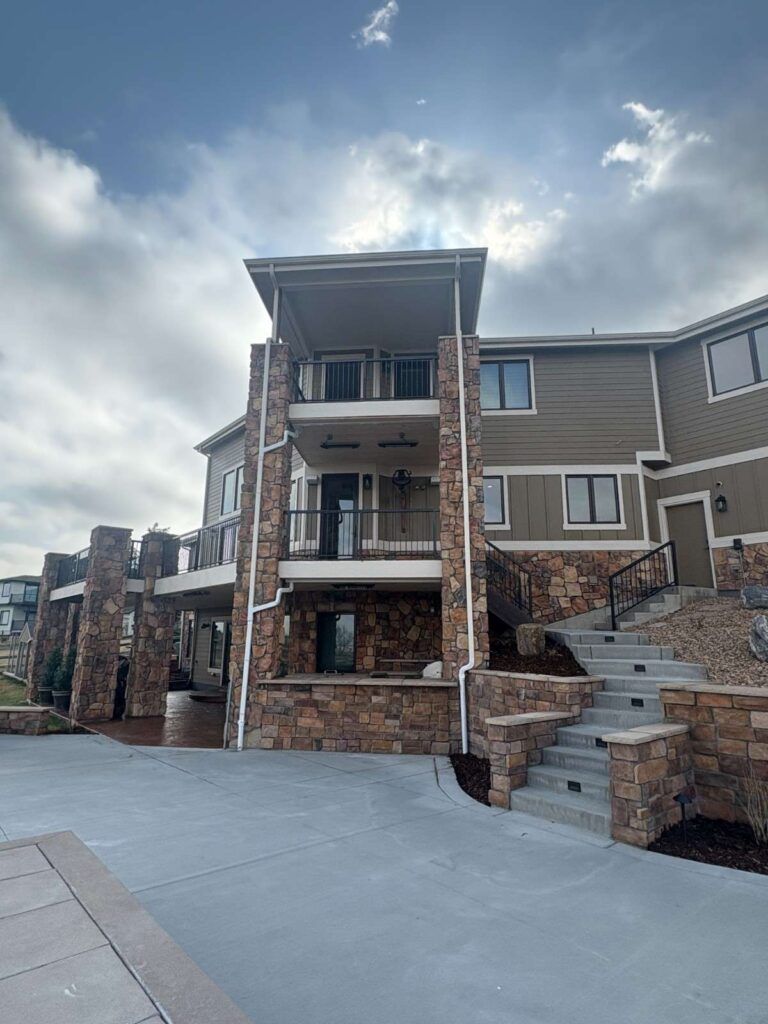 A multi-story house with stone pillars, a concrete patio, and a stone staircase under a cloudy sky.