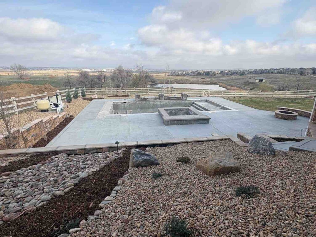 A stone patio under construction featuring an empty in-ground pool and fire pit, overlooking a rural landscape.