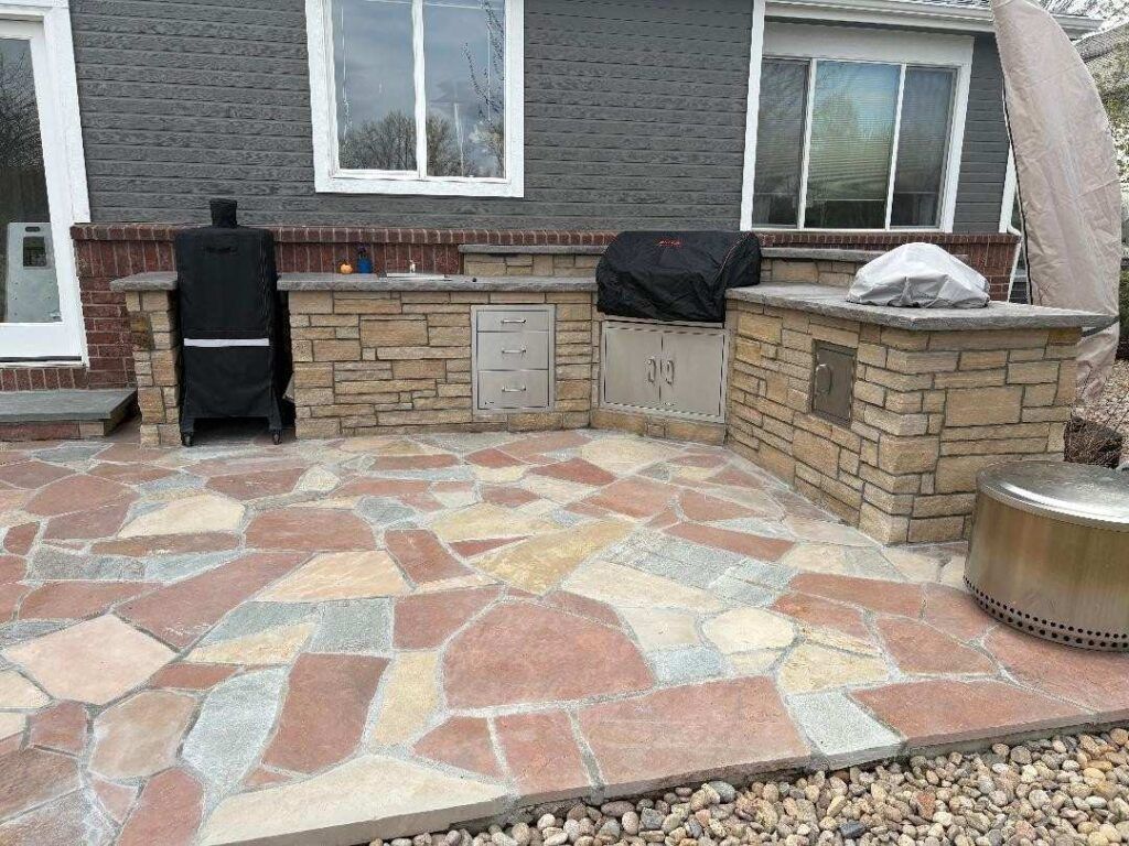 Outdoor patio with a stone kitchen island, grill, and smoker, finished with multicolored irregular flagstone paving.