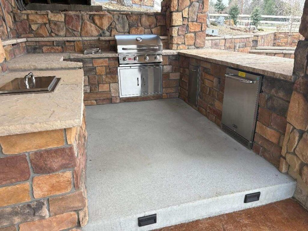 An outdoor stone kitchen area with a built-in stainless steel grill, a compact refrigerator, and a stone countertop.