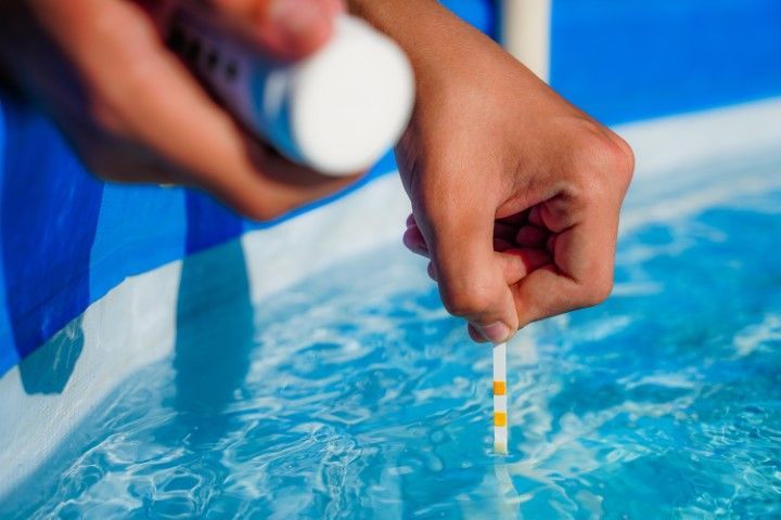 Hand holding a test strip over a blue swimming pool water sample