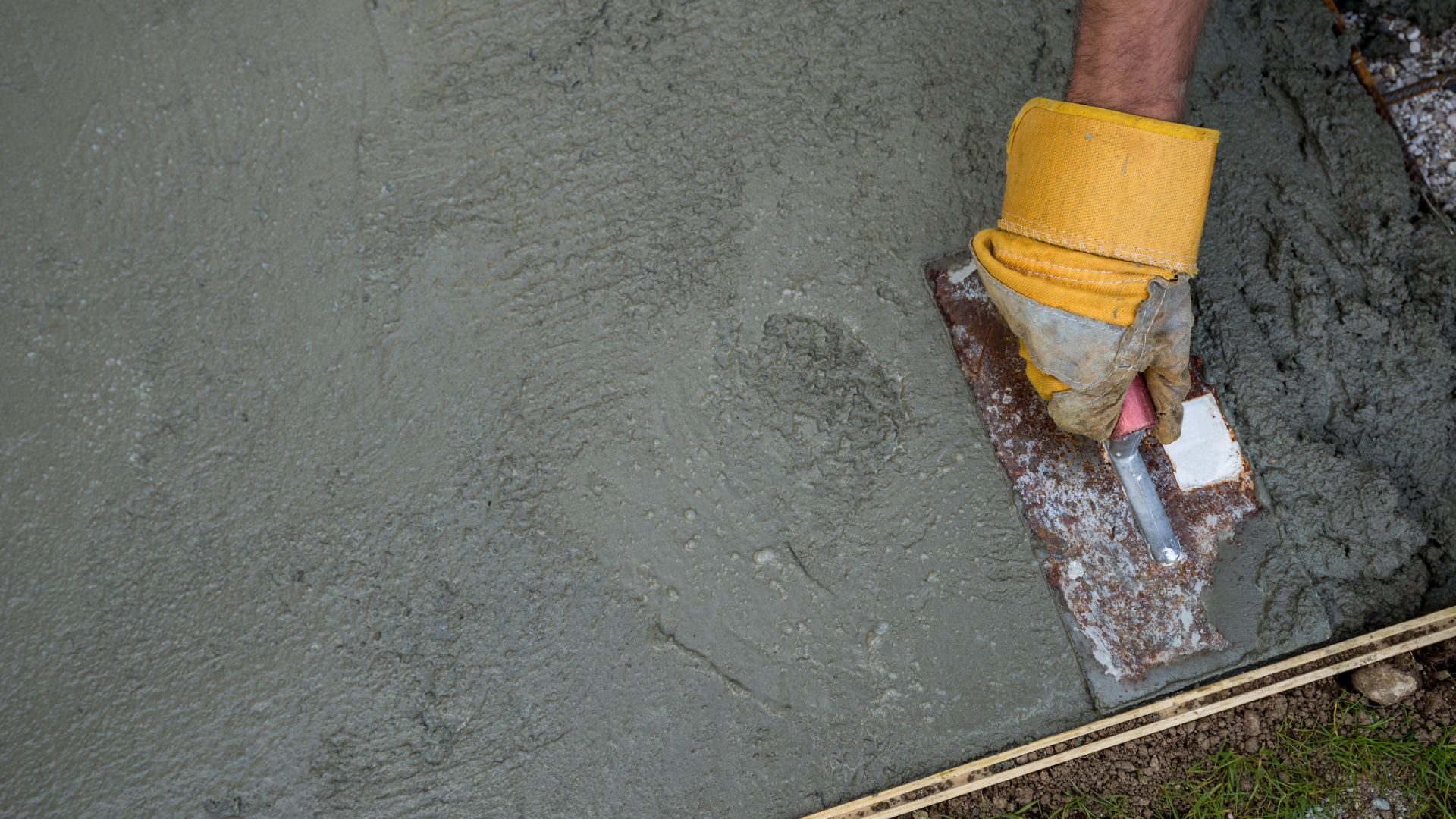 A trowel smooths wet gray concrete beside a yellow bucket and a rough wall edge.