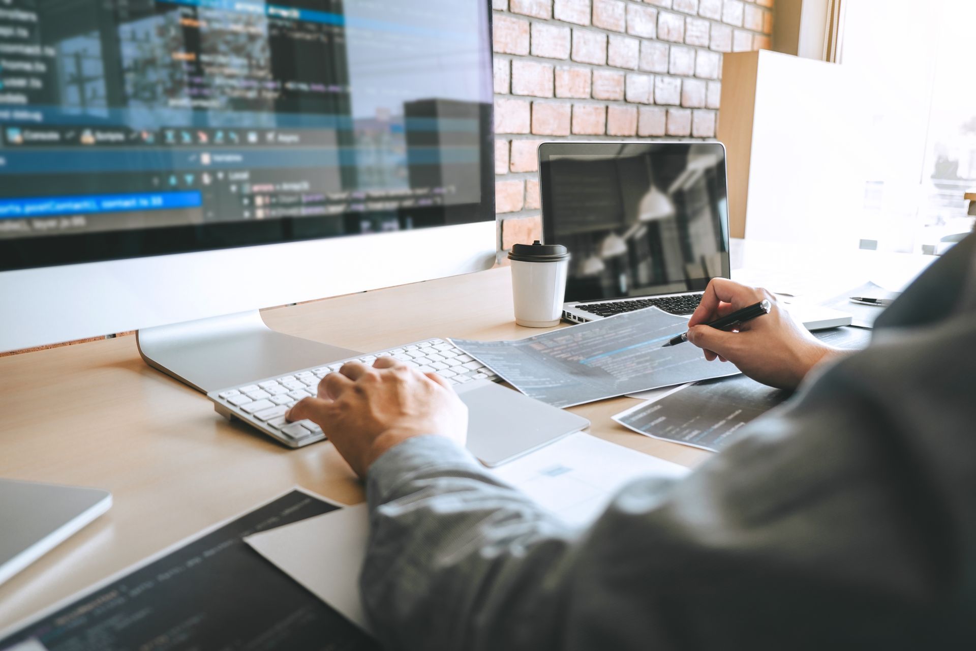 Person coding on a desktop computer with notes and a laptop in a modern office setting.