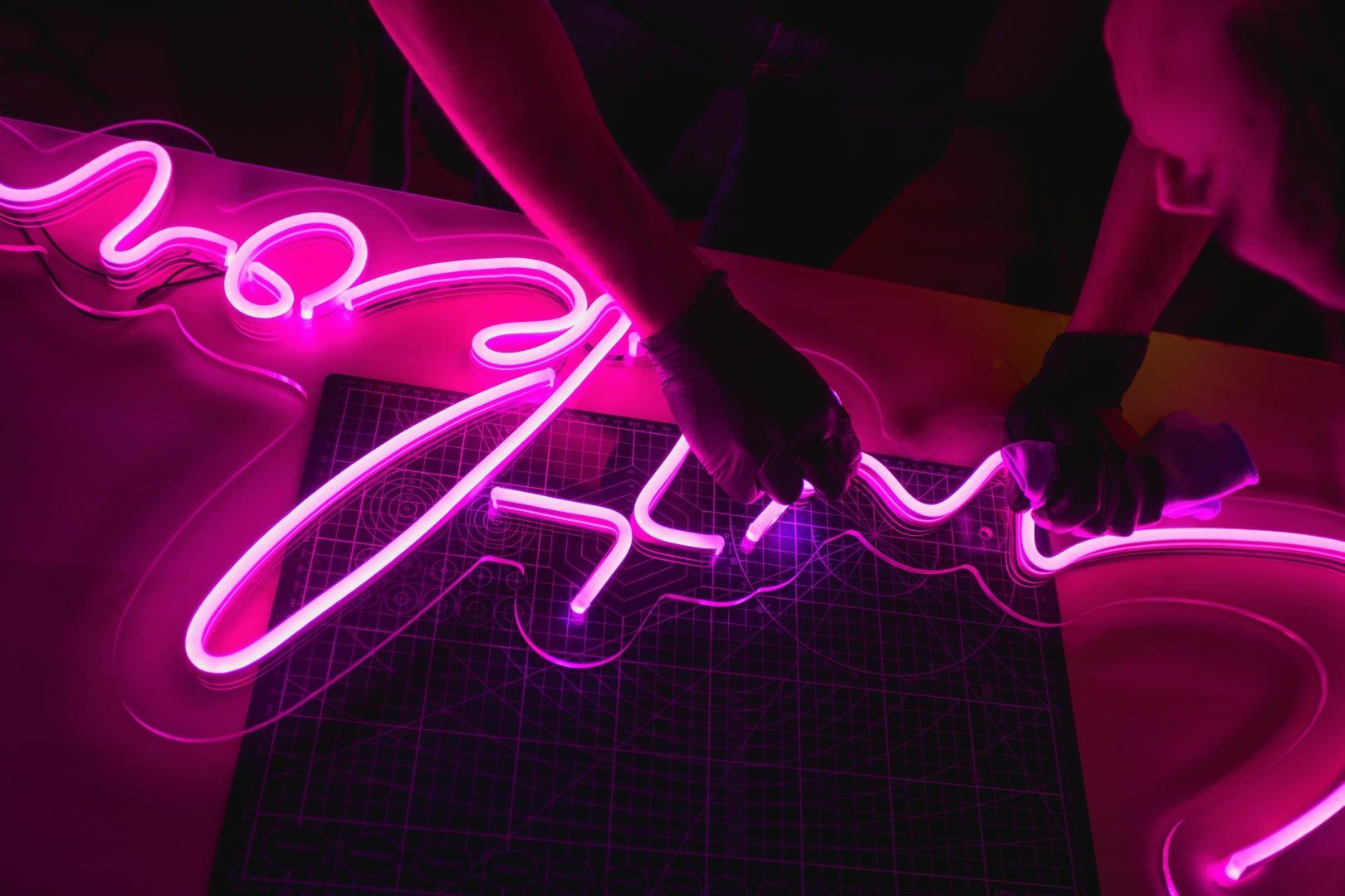 Hands working with a bright pink neon light, shaping it into a design on a dark surface.