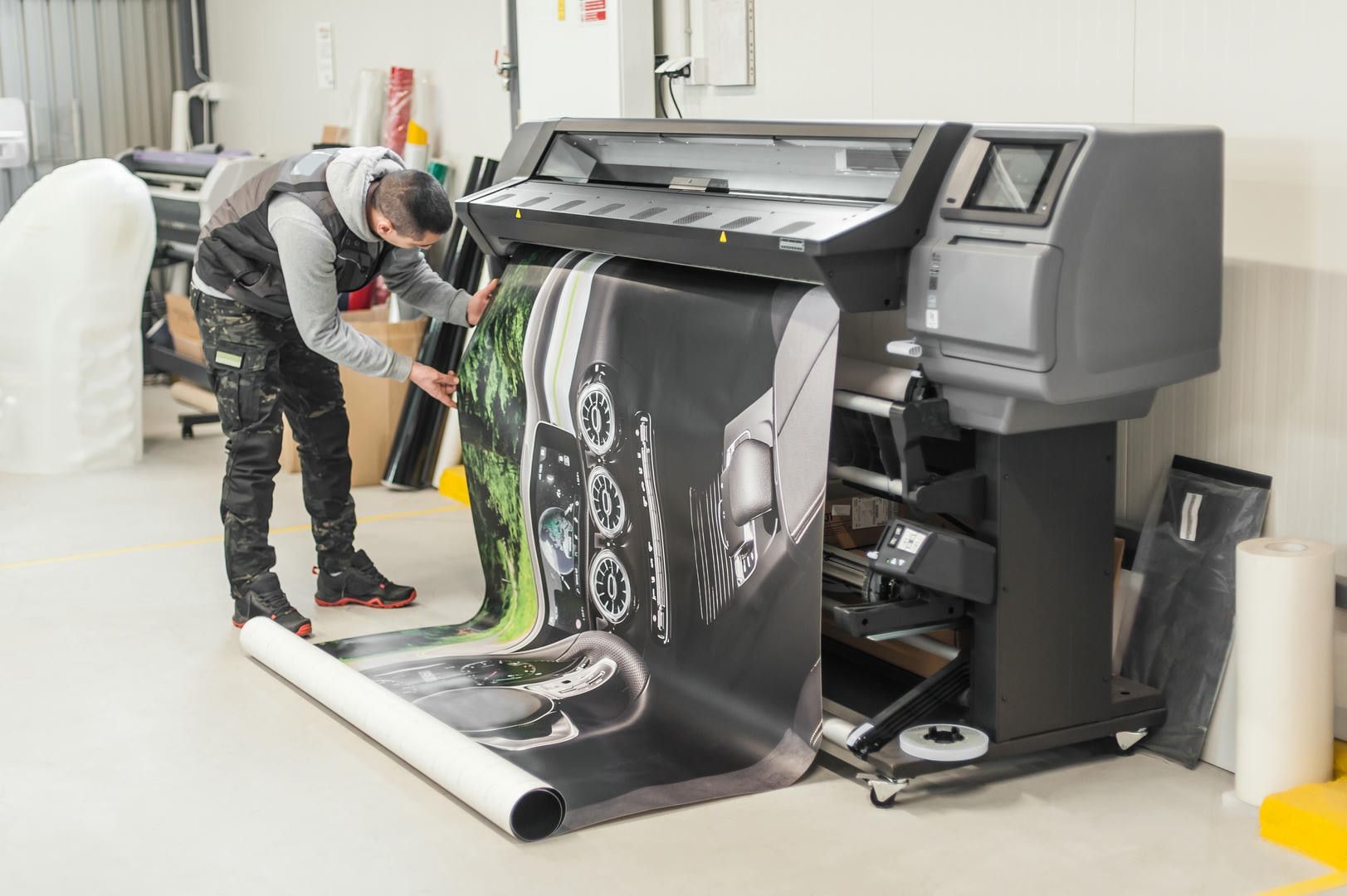 Man operating large-format printer, reviewing a printed graphic on roll of paper in a print shop.