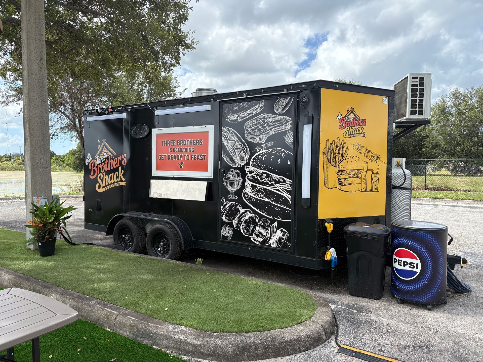 Black food trailer with burger art and Pepsi barrel, parked on grass beside a curb.