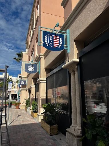 Blue and red storefront sign, outdoors on a building, with sidewalk, blue sky.