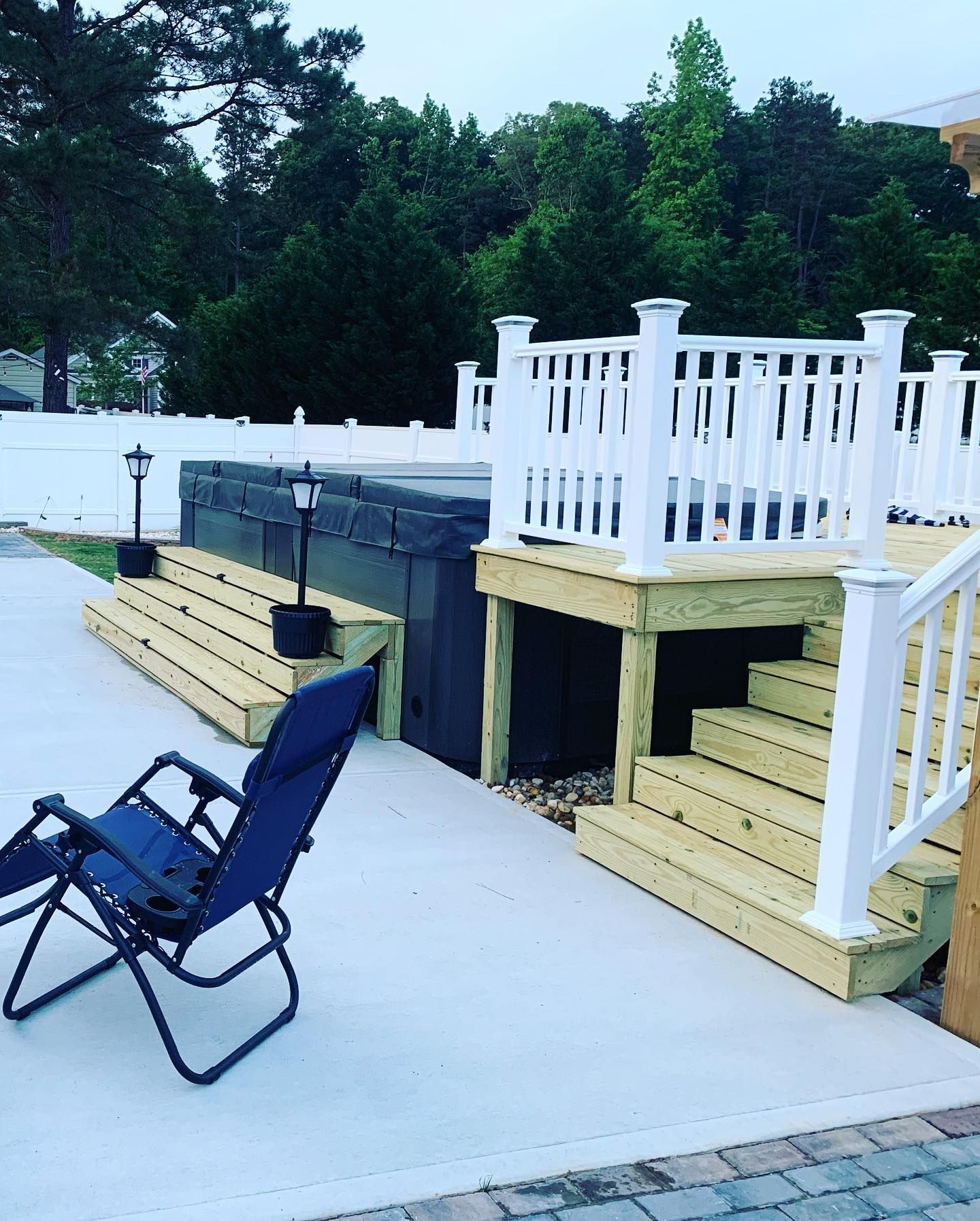 Hot tub on a wooden deck with stairs, white railing, and a blue chair on a concrete patio.