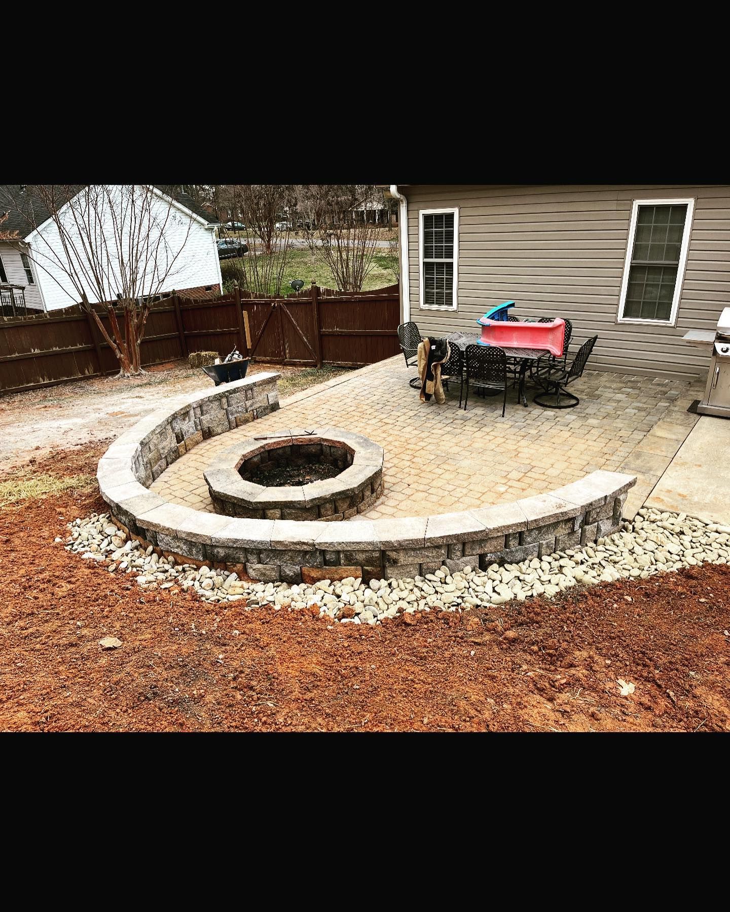 Curved stone retaining wall with fire pit and patio area in a backyard. Brown mulch and gravel surround.