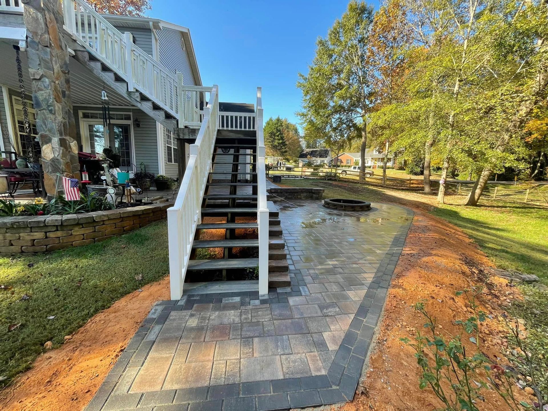 Wooden staircase from a two-story house leading to a paved patio and fire pit, next to grass and trees.