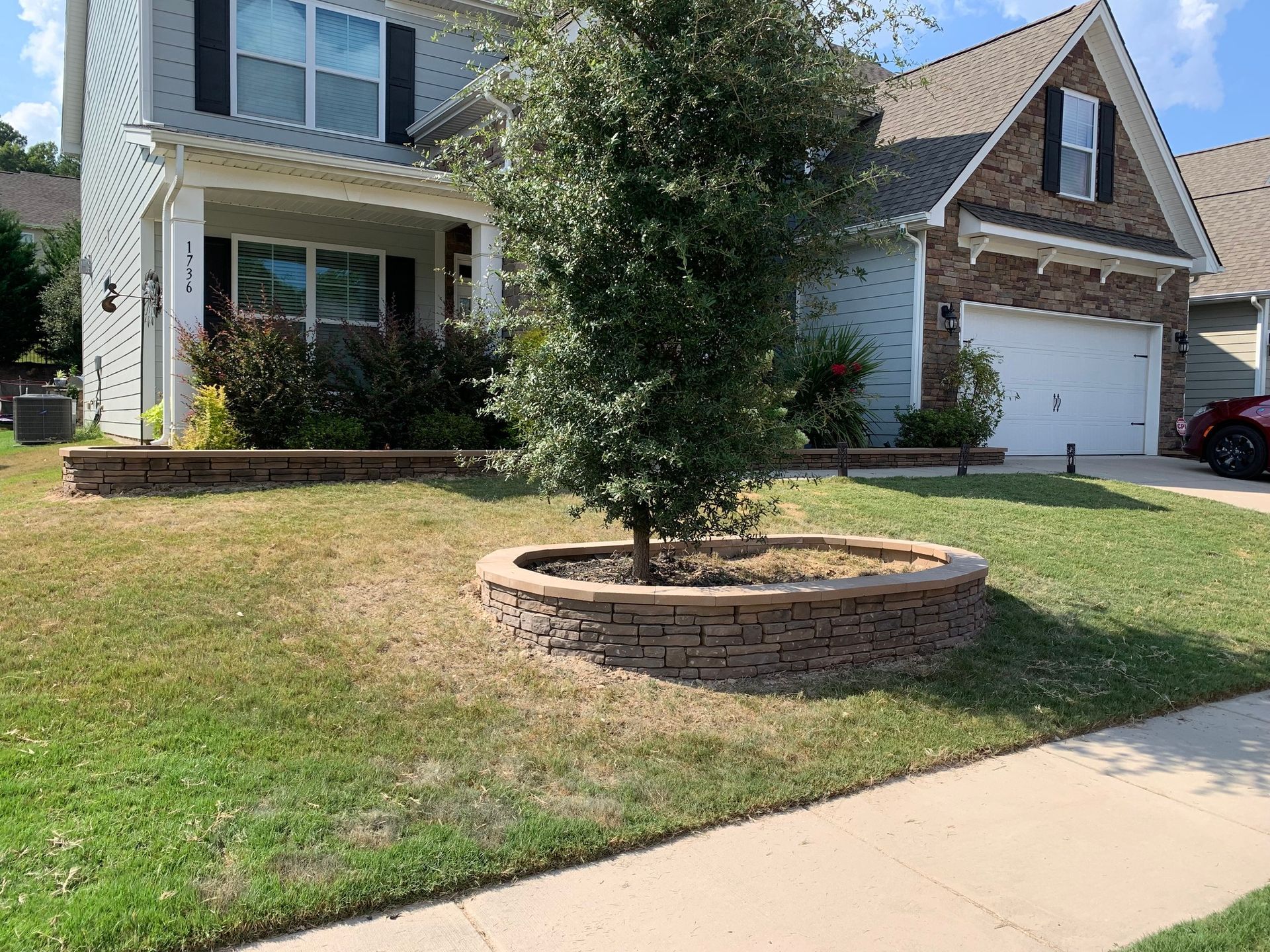 House with a lawn, tree ring, and retaining walls. Overcast sky and a sidewalk in front.