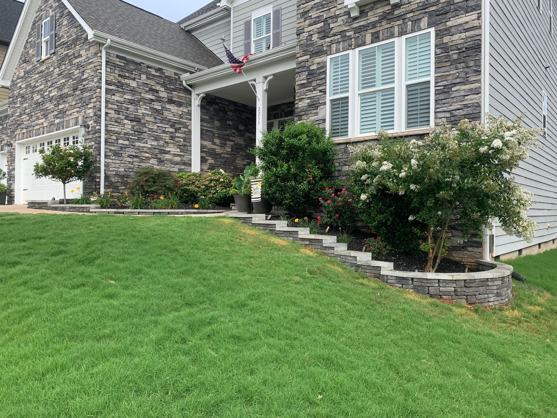 House with stone facade, green lawn, retaining wall with shrubs.