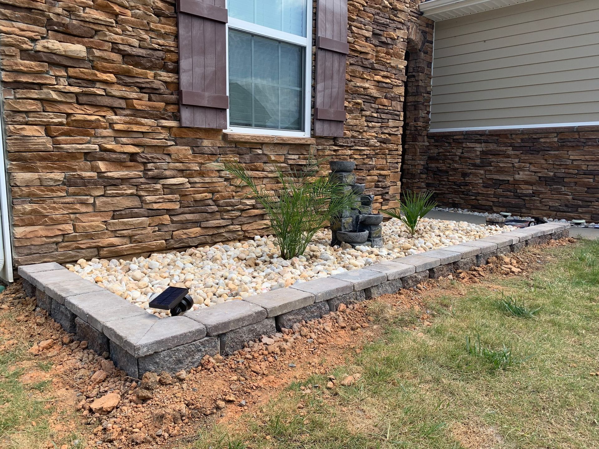 Stone-walled house with a garden bed, plants, rocks, and a small statue next to the house.