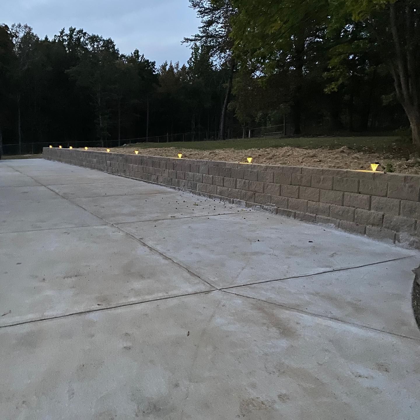 A concrete driveway next to a low brick wall with lights, set against a wooded background.