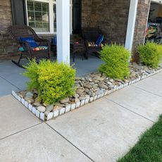 A front porch with yellow-green bushes, pebbles, and two wicker rocking chairs.
